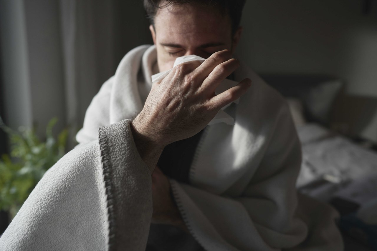 Man sitting up in bed with a blanket, blowing his nose