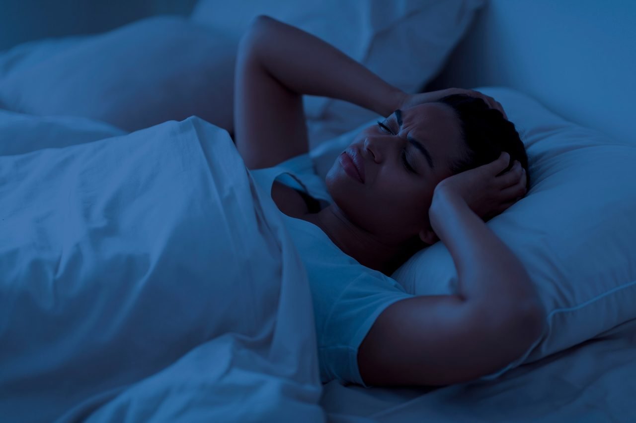 Nighttime clenching often shows up as tension and headaches Woman lying awake in bed at night pressing her hands against her head in discomfort