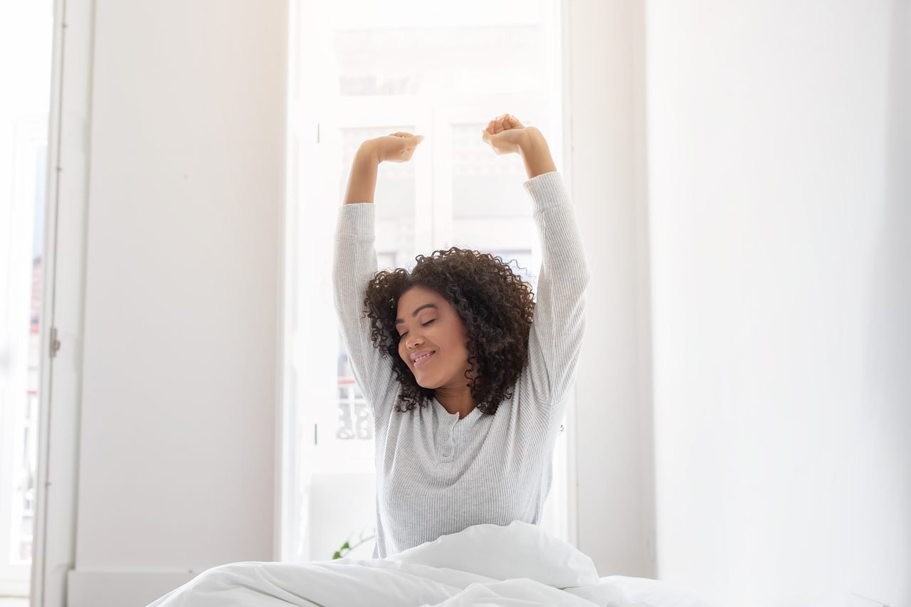 Waking up without jaw soreness is the goal of a facial pain evaluation Woman sitting up in bed and stretching her arms overhead after waking up